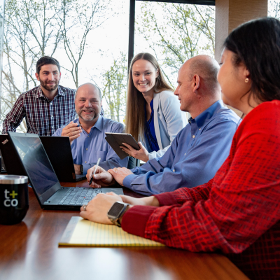 Five employees surround a conference table, smiling, while on computers and tablets.