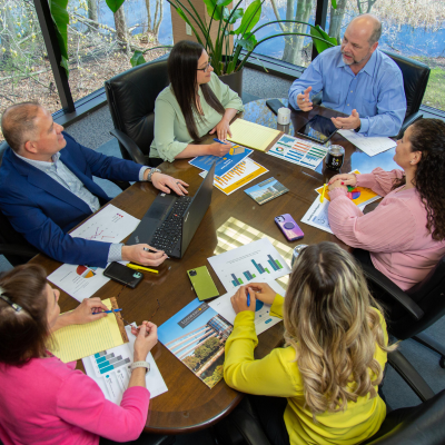 Six employees discuss at a large conference table with accounting charts, pamphlets, and information scattered on the table.