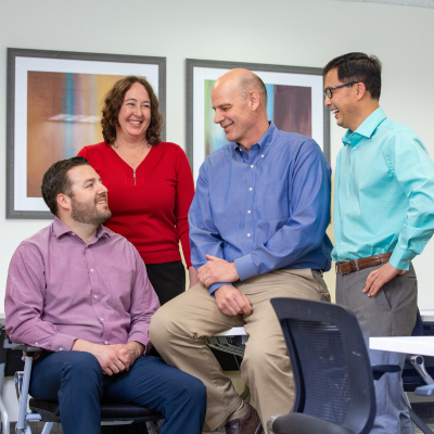 Four employees standing together and smiling in a conference room, one sits on the edge of a table, two stand, and one sits in an office chair.
