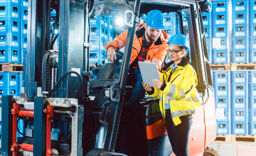 A distribution worker shows a clipboard to another distribution worker who is sat in a forklift, both in front of a wall stacked high with blue boxes.