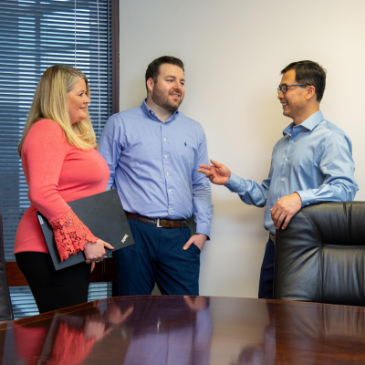 Three accounting professionals smile while standing near a conference table, one professional gestures with his hand while the other two face him.