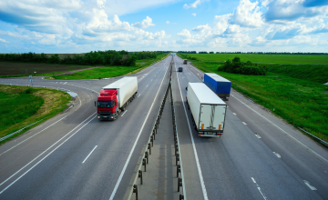 A two way highway surrounded by bright green fields and blue cloudy skies, has two trucks going North and one truck going South.