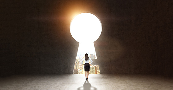 A business woman stands in an empty room in front of a large key hole, which reveals sunlight light and a city skyline.