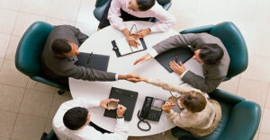 A Bird's eye view of five business professionals sat at a round circle shaking each other's hands.
