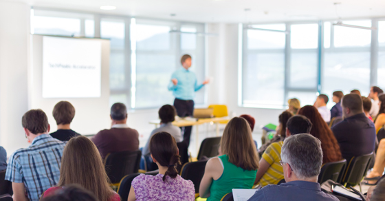 An image of a business training session, where a man stands at the head of a classroom in front of a projector speaking to a room full of individual's sitting in chairs.