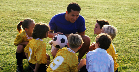 A man crouches on a field of grass with a soccer ball in his hands as he speaks to six little soccer players.