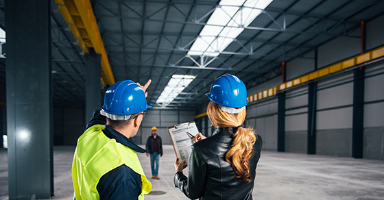 A woman points to a clipboard and stands next to a man as he points to the ceiling of the empty warehouse they are inside. Both wear a a hard hat as a man in the background walks toward them.