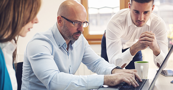 Three business professionals sit looking at a laptop screen; a man types on the computer, while another mean leans over his right side and a women sits to his left viewing the computer.