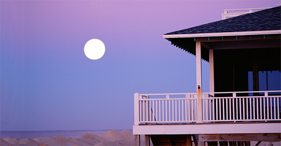 A beach house with a wrap around porch during a blue and pink sunset, with a full moon in the distance.