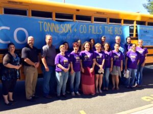 Tonneson employees stand in front of a yellow school bus with a banner that states "Tonneson + Co Fills The Bus" and are wearing purple, Caties Closet shirts.