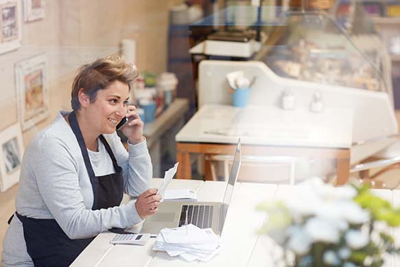 A smiling woman, standing in a restaurant, wears an apron while on the phone holding a restaurant ticket.