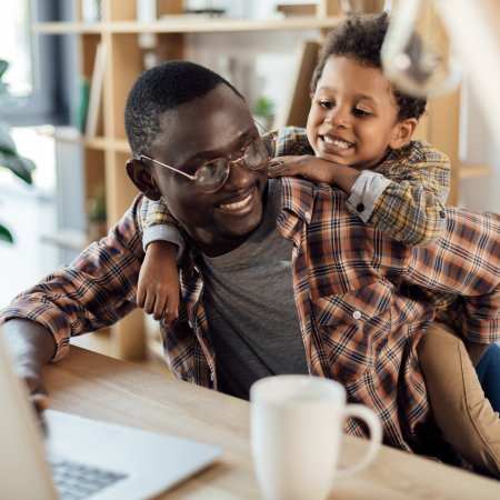 Man sitting at desk with computer with his son on his back